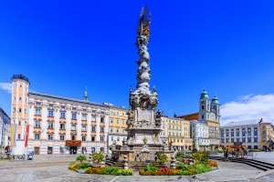 Barocke Dreifaltigkeitssäule auf einem Platz mit historischen Gebäuden im Hintergrund.