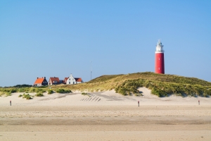 Roter Leuchtturm auf Düne mit Häusern und blauem Himmel im Hintergrund.