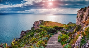 Sonnenuntergang über einer Küstenlandschaft mit klarem Himmel und Meer. Ein Pfad schlängelt sich durch die grüne Vegetation entlang einer felsigen Klippe.