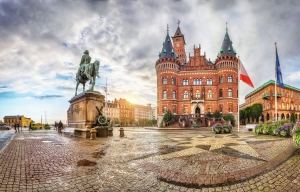 Rotes historisches Gebäude mit Turm und Reiterstatue auf Stadtplatz bei bewölktem Himmel.