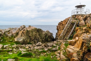 Klippe mit kleinem Leuchtturm und Treppe am Meer bei bewölktem Himmel.