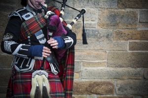 Person in traditioneller schottischer Kleidung spielt Dudelsack vor einer Steinmauer.