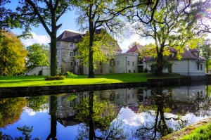 Ein historisches Gebäude, möglicherweise ein Schloss oder Herrenhaus, liegt inmitten von Bäumen. Es steht an einem ruhigen Gewässer, das die Struktur und die umliegende Vegetation klar spiegelt. Das Wetter ist sonnig und der Himmel ist teilweise bewölkt.