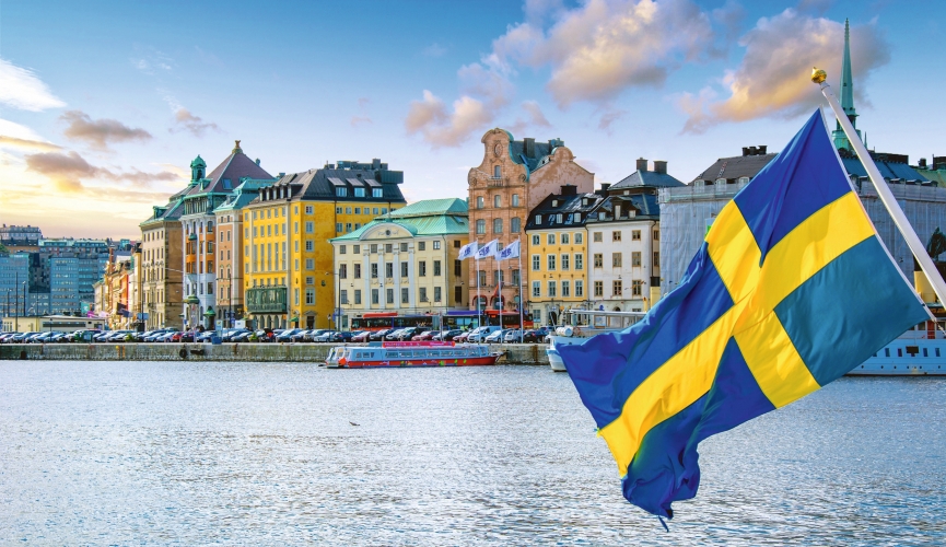 Schwedische Flagge weht vor bunten Gebäuden am Wasser.