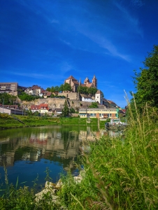 Burg auf einem Hügel mit Fluss im Vordergrund an einem sonnigen Tag.