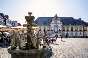 Das Bild zeigt das Rathaus von Lippstadt in Nordrhein-Westfalen, Deutschland, mit einer Pflasterung im Vordergrund und Menschen, die draußen sitzen.
