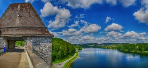 Alter Staudamm an der Möhnesee-Talsperre in Nordrhein-Westfalen, Deutschland, umgeben von bewaldeten Ufern und einem klaren, blauen Himmel mit weißen Wolken.