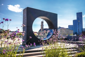 Skulptur mit kreisförmiger Öffnung, umgeben von lila Blumen und Stadtgebäuden im Hintergrund.