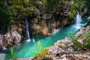 Wasserfall fließt in smaragdgrünen Fluss in bewaldeter Felslandschaft.