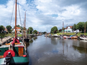 Segelboote an einem Fluss mit Uferpromenade und Häusern im Hintergrund unter bewölktem Himmel