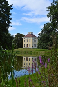 Gelbes Herrenhaus spiegelt sich in einem Teich, umgeben von grünen Bäumen und Pflanzen.