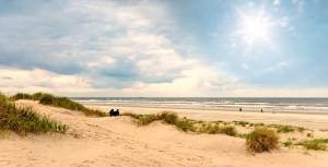 Strand mit Dünen, grasbewachsen, sonniger Himmel, ruhiges Meer im Hintergrund.