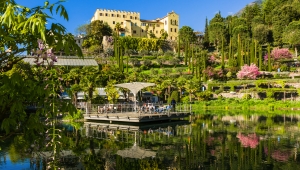 Gelbes Schloss inmitten grüner Gärten und Blüten, mit See im Vordergrund.