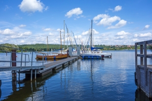 Segelboote an einem Steg auf einem ruhigen See unter blauem Himmel mit weißen Wolken, im Hintergrund eine Landschaft mit grünen Hügeln und vereinzelt Häusern.