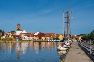 Segelboote, die an einem Kai in einem ruhigen Hafen festgemacht sind, mit einer Altstadt im Hintergrund, die von bunten Gebäuden und einem markanten Kirchturm geprägt ist, unter einem klaren blauen Himmel.
