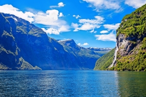 Blick auf einen Fjord mit Wasserfall, umgeben von bewaldeten Bergen unter blauem Himmel