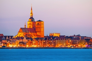Blick auf die historische Altstadt von Stralsund bei Sonnenuntergang mit beleuchteten Gebäuden und dem Wasser der Ostsee im Vordergrund.