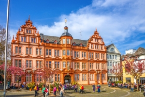 Historisches Gebäude mit roten Fensterläden und blühenden Bäumen davor.
