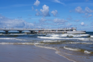Wellen am Strand vor einer langen Seebrücke mit zahlreichen Spaziergängern.