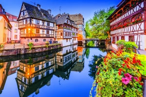 Fachwerkhäuser am Wasser mit Blumen im Vordergrund und spiegelnder Wasserfläche.