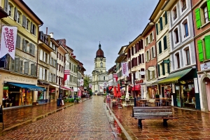 Pflasterstraße in einer belebten Altstadt mit Geschäften und Kirche im Hintergrund.