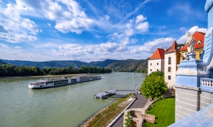 Flusskreuzfahrtschiff auf der Donau vor Schloss und Hügeln unter blauem Himmel.