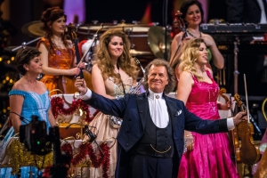 Mann im Frack dirigiert Orchester beim Konzert, Musikerinnen in Ballkleidern im Hintergrund.