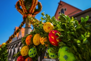 Ein traditionell mit bunten Ostereiern und grünem Laub geschmückter Osterbrunnen vor einem blauen Himmel und einem Gebäude im Hintergrund.