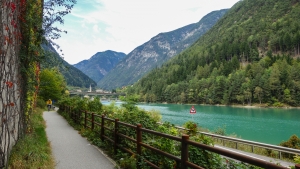 Radweg entlang eines Sees mit Bergen und Wald im Hintergrund.