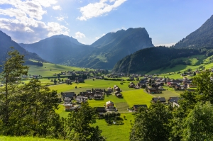 Alpendorf in grüner Berglandschaft mit Häusern und umliegenden Bergen im Sonnenlicht.