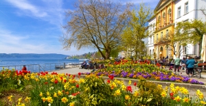 Park am See mit Blumenbeeten, Bäumen und Spaziergängern bei sonnigem Wetter.