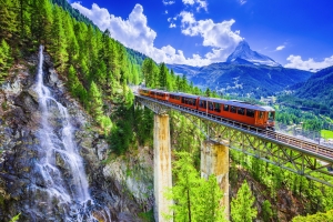 Roter Zug auf Brücke vor Wasserfall und Berglandschaft mit blauem Himmel.