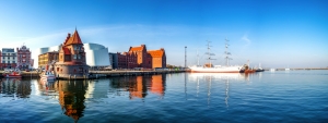 Panoramafoto eines Hafens mit historischen Gebäuden und einem großen Segelschiff am Wasser bei klarem Himmel.