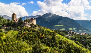 Burg auf Hügel mit Berglandschaft im Hintergrund, umgeben von grüner Natur.