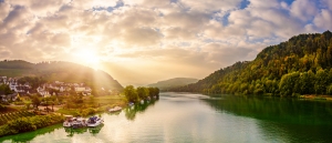 Flusslandschaft mit Bergen, Häusern und Sonnenaufgang im Hintergrund