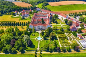 Luftaufnahme eines großen Schlosses mit Gartenanlage und umgebenden Feldern und Bäumen.