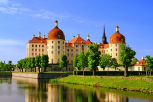 Schloss mit Türmen und rotem Dach am Wasser unter blauem Himmel.