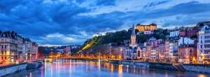 Stadtansicht von Lyon bei Dämmerung mit erleuchteten Gebäuden am Fluss.