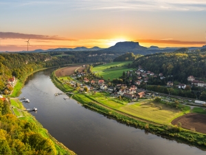Landschaft mit Fluss, Windrad, Dorf und Sonnenuntergang in hügeliger Gegend.