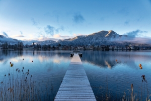 Steg führt über einen ruhigen See zu schneebedeckten Bergen im Hintergrund.