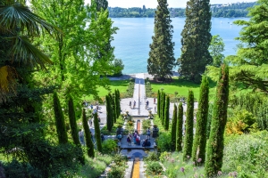 Gartenanlage mit hohen Zypressen und Blick auf einen See im Hintergrund.
