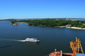Fluss Świna bei Swinemünde mit einem Schiff auf dem Wasser, umgeben von grüner Landschaft und einem sandigen Ufer. Im Hintergrund sind die Inseln Usedom und Wollin sowie weitere Gebäude entlang der Ostseeküste zu sehen.