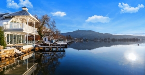 See mit Uferhäusern und schneebedeckten Bergen im Hintergrund bei klarem Himmel.