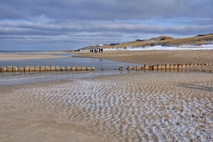 Sandstrand mit Holzpfählen, dahinter Dünen und Wolkenhimmel.