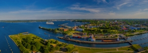Panorama-Luftbild der Wasserstraße Nord-Ostsee-Kanal mit der Schleusenanlage Holtenau, inklusive eines Kreuzfahrtschiffs und mehreren Fracht- und Transportschiffen in der Nähe, umgeben von Landschaft und Küstensiedlungen.