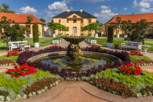Historischer Garten mit Springbrunnen, umgeben von Blumen und Gebäuden im Barockstil.