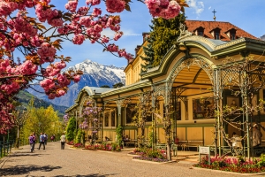 Historisches Gebäude mit blühenden Bäumen und schneebedeckten Bergen im Hintergrund.