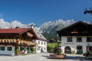 Dorfplatz mit traditionellen Häusern und Bergen im Hintergrund bei klarem Himmel.