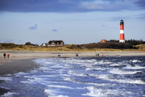 Strand mit Leuchtturm und Spaziergängern bei Wellengang und Wolkenhimmel.