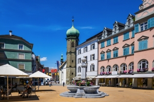 Stadtplatz mit historischen Gebäuden und einem zentralen Brunnen bei klarem Himmel.